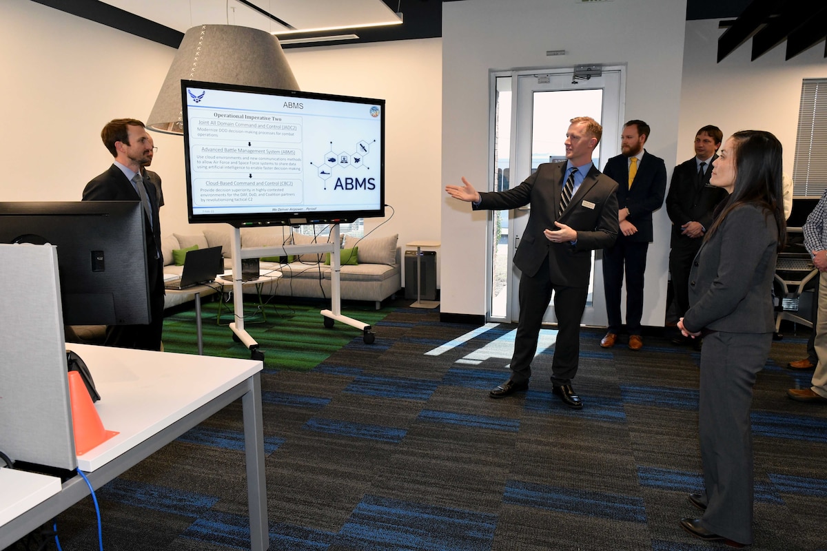 Photo shows group of people standing in room looking at large tv screen