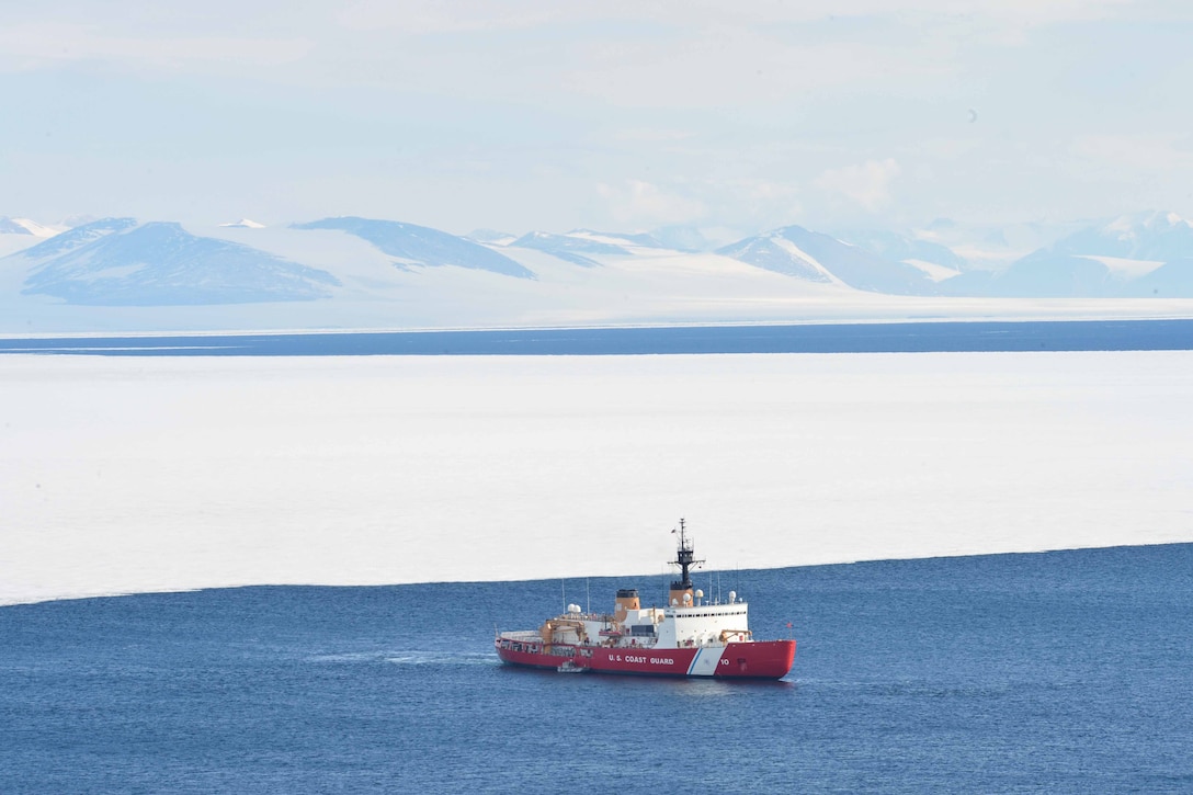 A Coast Guard ship travels across the water with snowy mountains in the background.