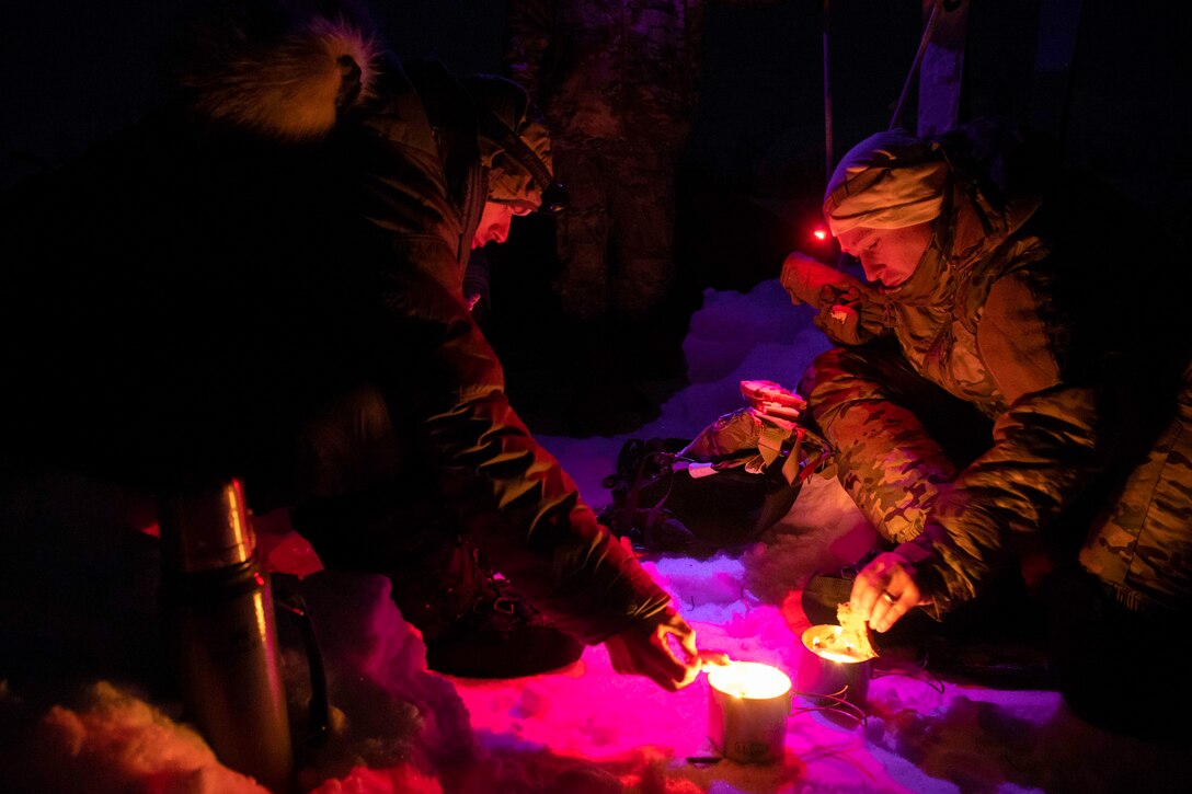 One airman dips bread into a small pot as another airman holds onto the lid at night.