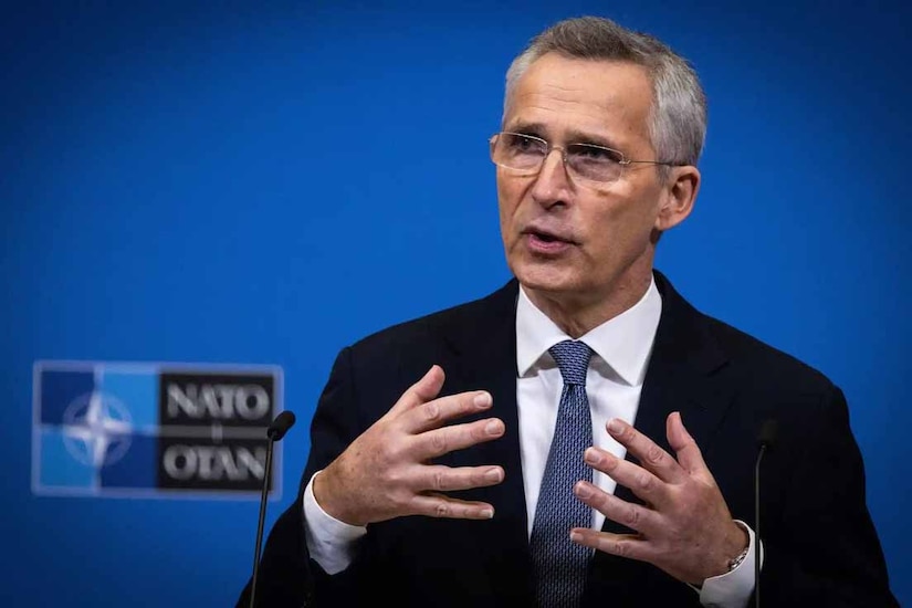 A man gestures as he speaks. The sign behind him indicates that he is at NATO headquarters.