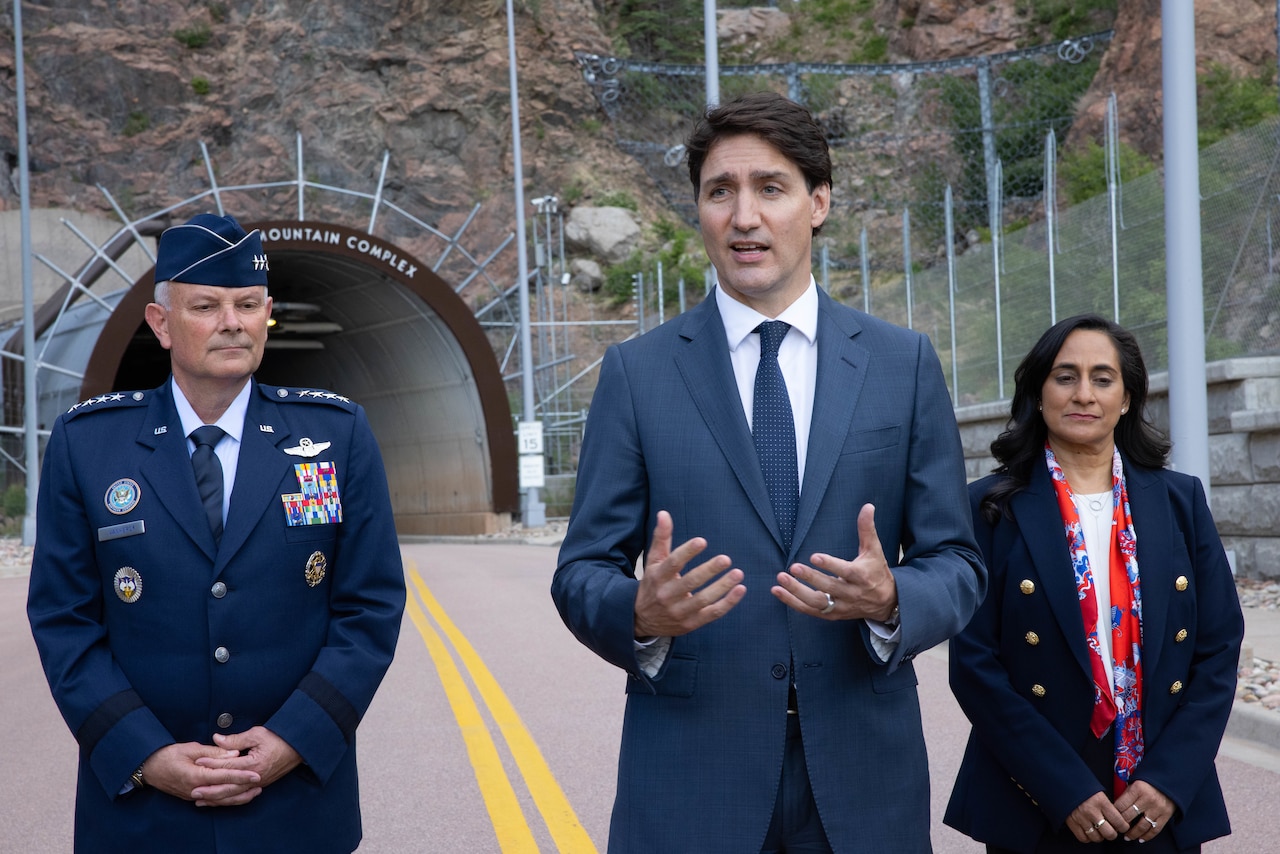 Two men, one in a military uniform, and a woman stand outside a tunnel entrance at the side of a mountain.