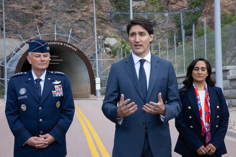 Two men, one in a military uniform, and a woman stand outside a tunnel entrance at the side of a mountain.