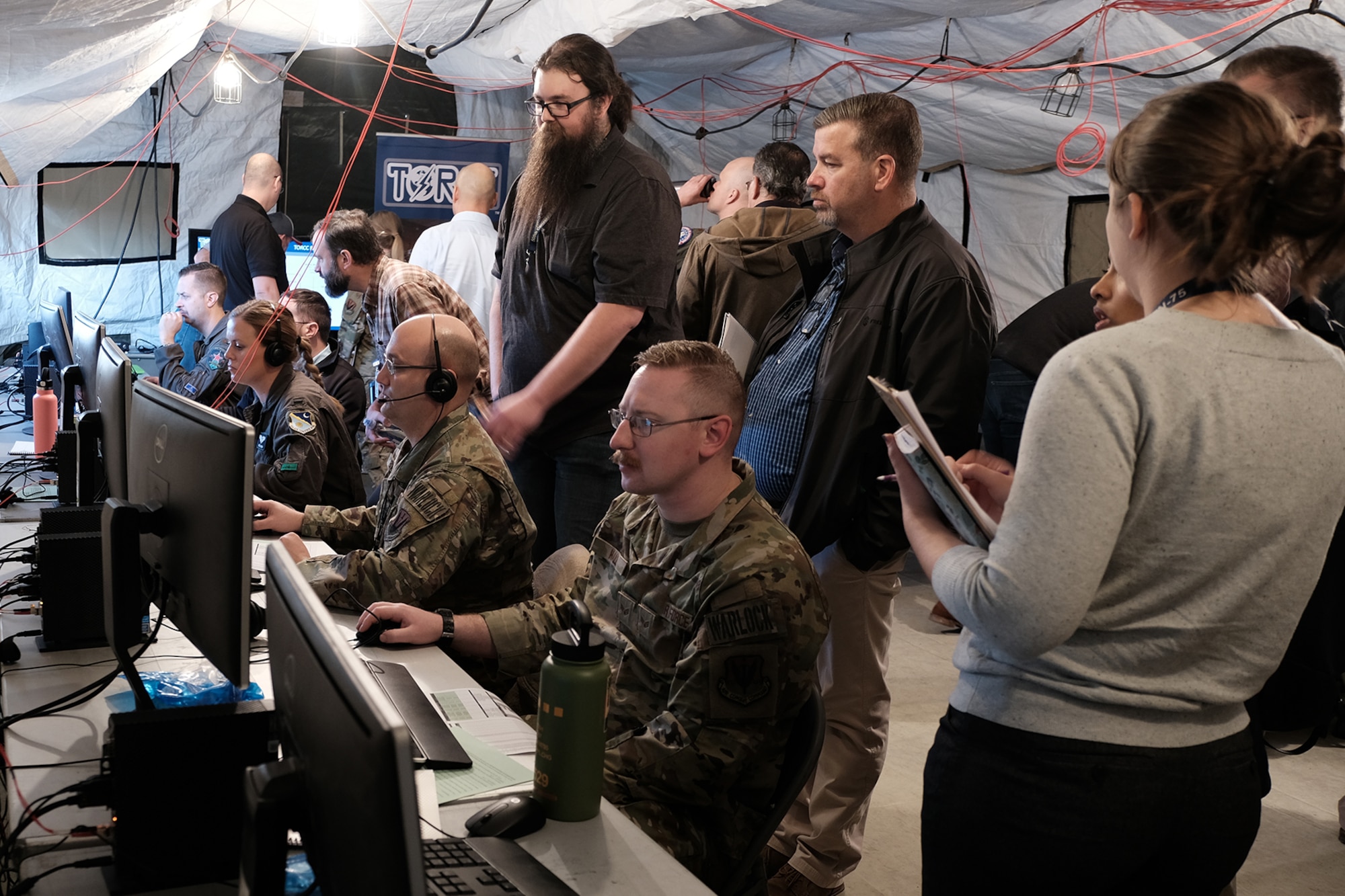 photo of U.S. Air Force military members work on computers in a tent while civilians stand and watch