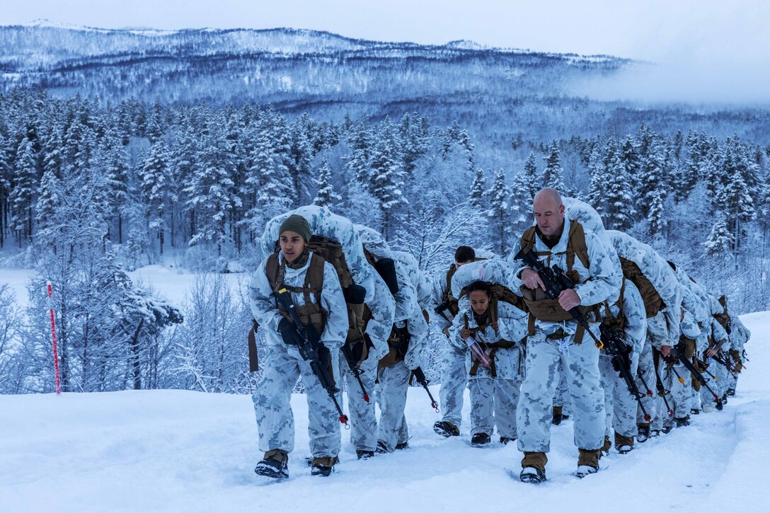 A group of Marines hike up a mountain in the snow with rucks on their backs.