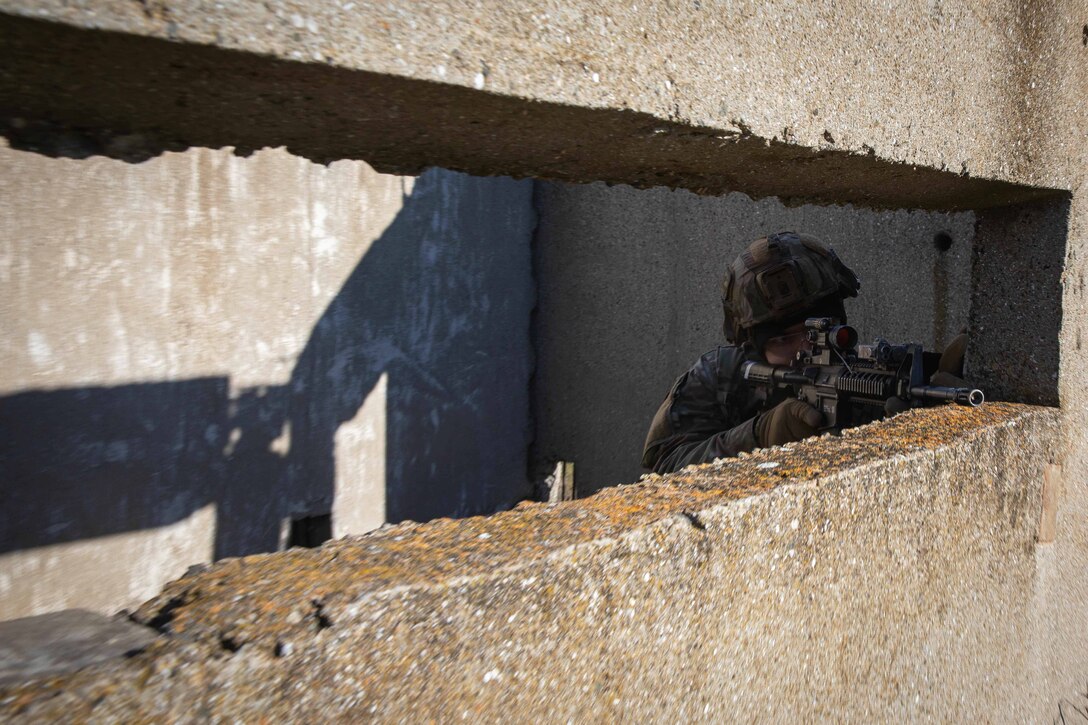 A soldier holding a weapon looks through the scope from a bunker.