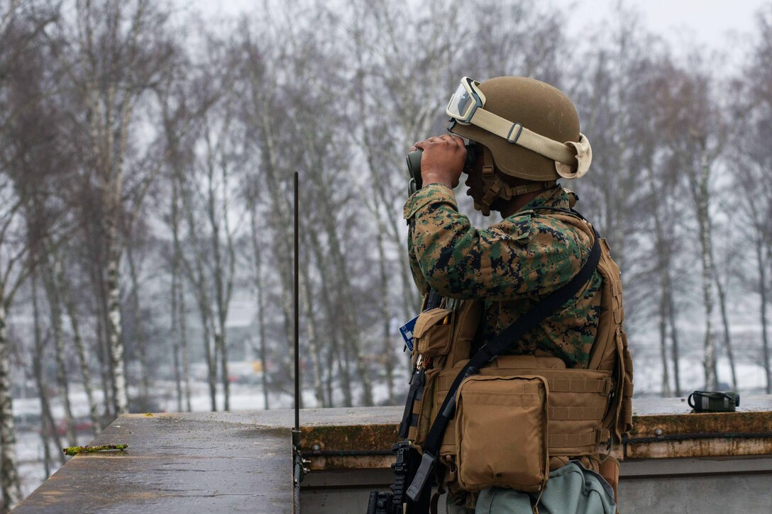 A soldier wearing full combat gear looks through binoculars during a security drill.