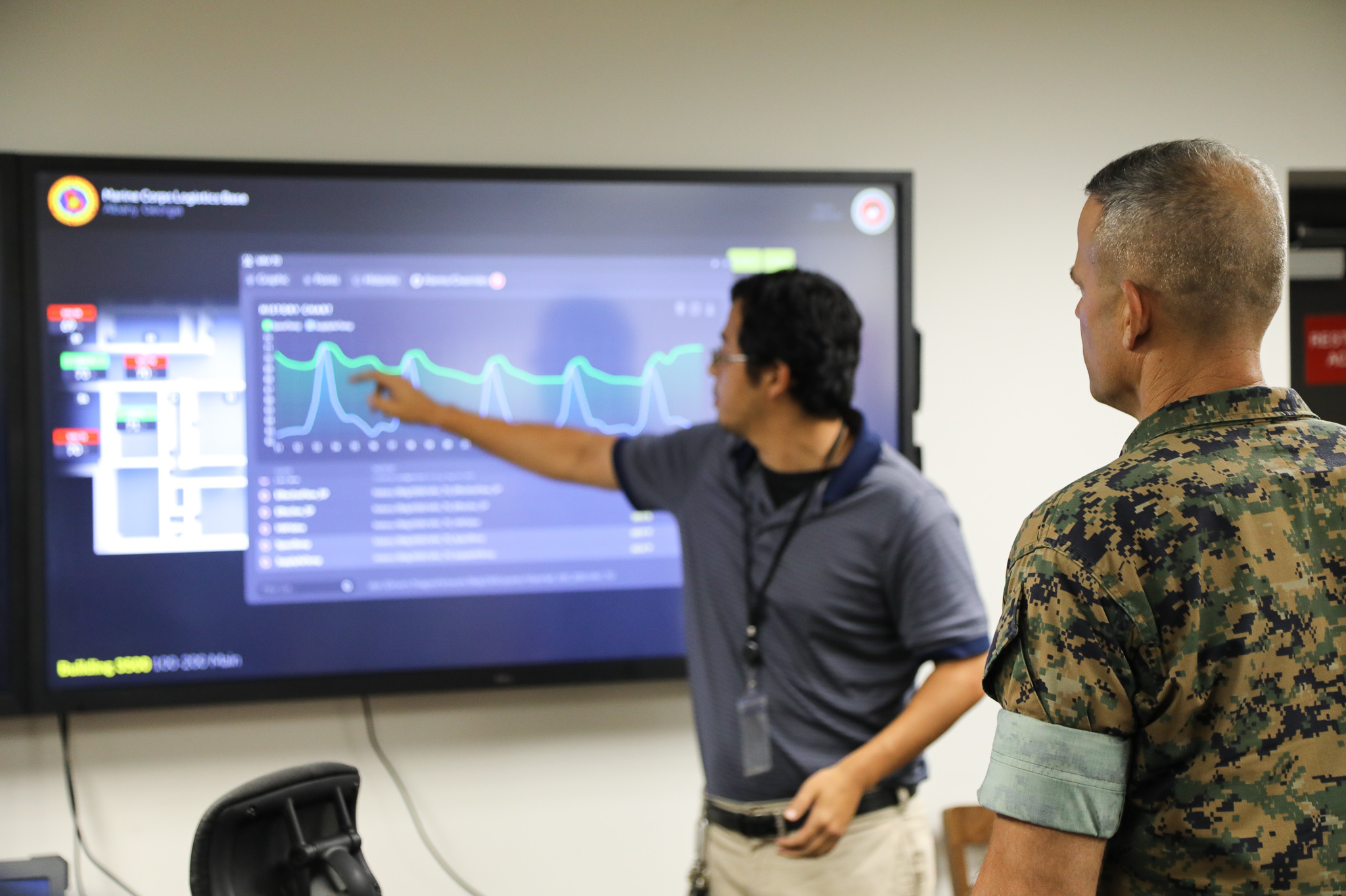 A man pointing to a graph on a TV screen with a Marine listening in the foreground.