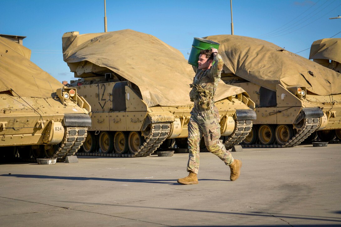 A soldier carrying a green bucket walks near parked armored vehicles covered in beige tarps.