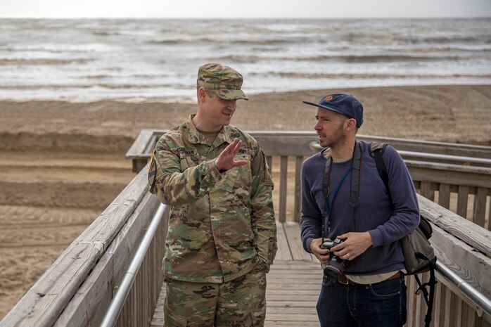 two people stand on a wooden walkway leading to an ocean beach talking to each other.
