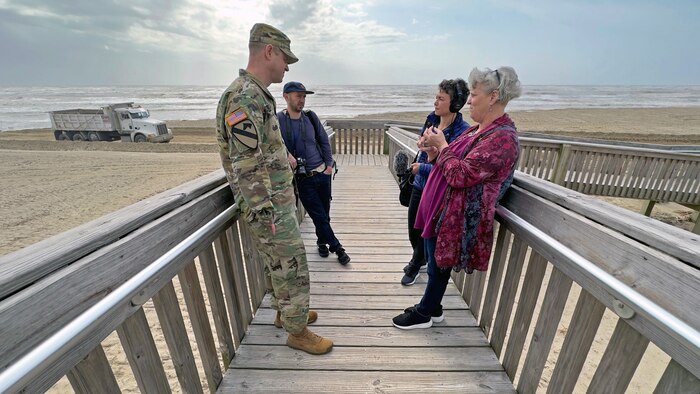 four people stand on a wooden walkway leading to an ocean beach talking to each other. A dump truck drives by in the background.