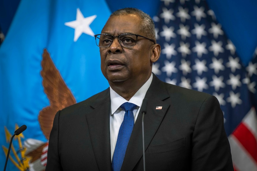 A man stands behind two microphones. Flags are in the background.