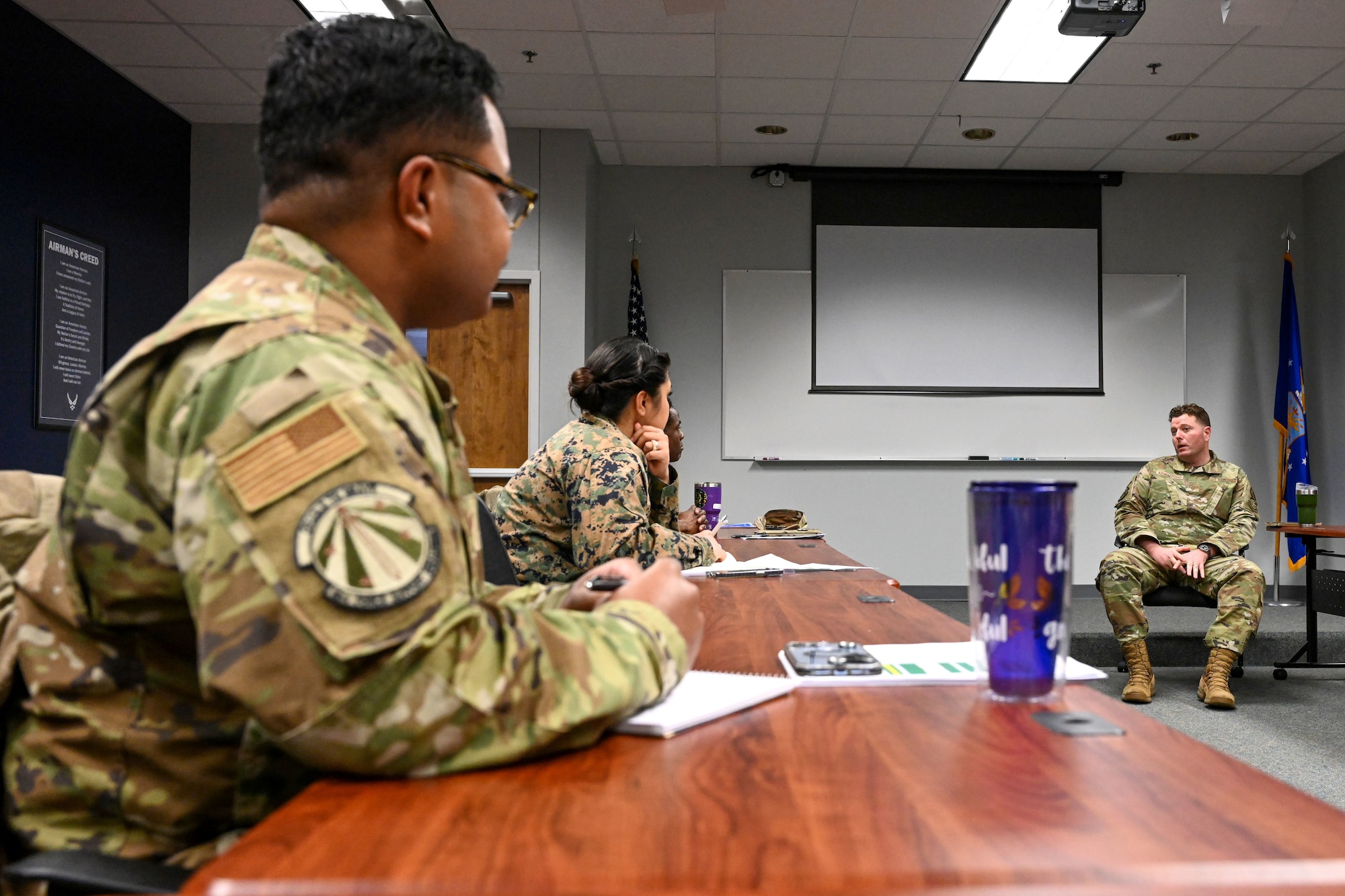 Two people sit at a table, listening to a man speak.