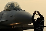 A man stands in front of a fighter jet and uses hand signals to direct the pilot to park the aircraft.