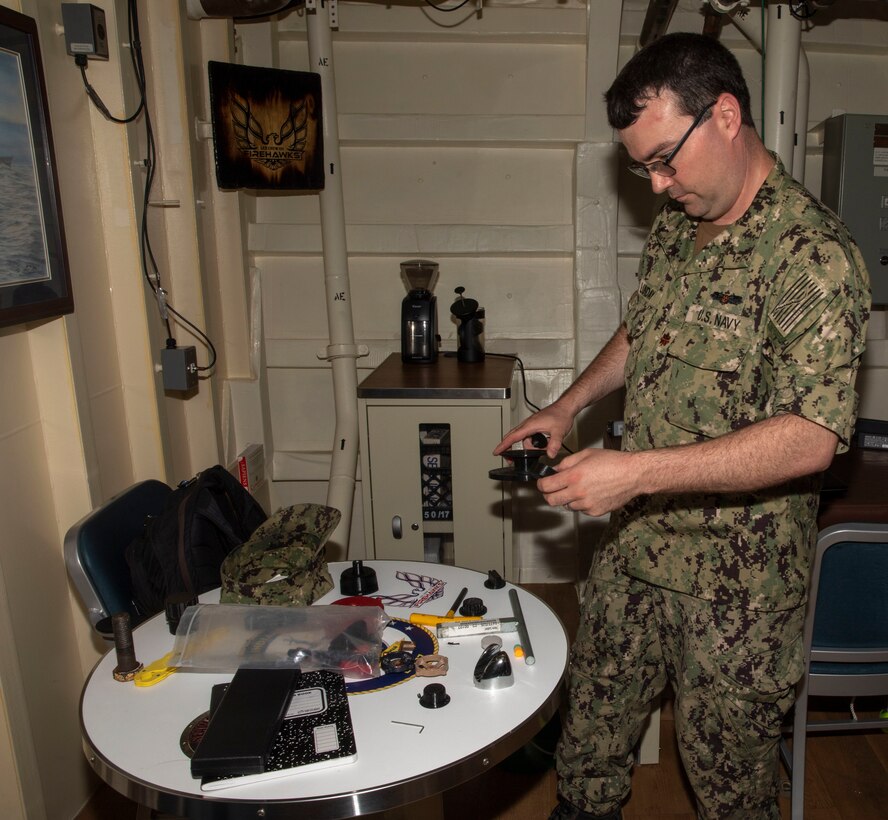 Man in camo uniform stands in front of a table with miscellaneous items