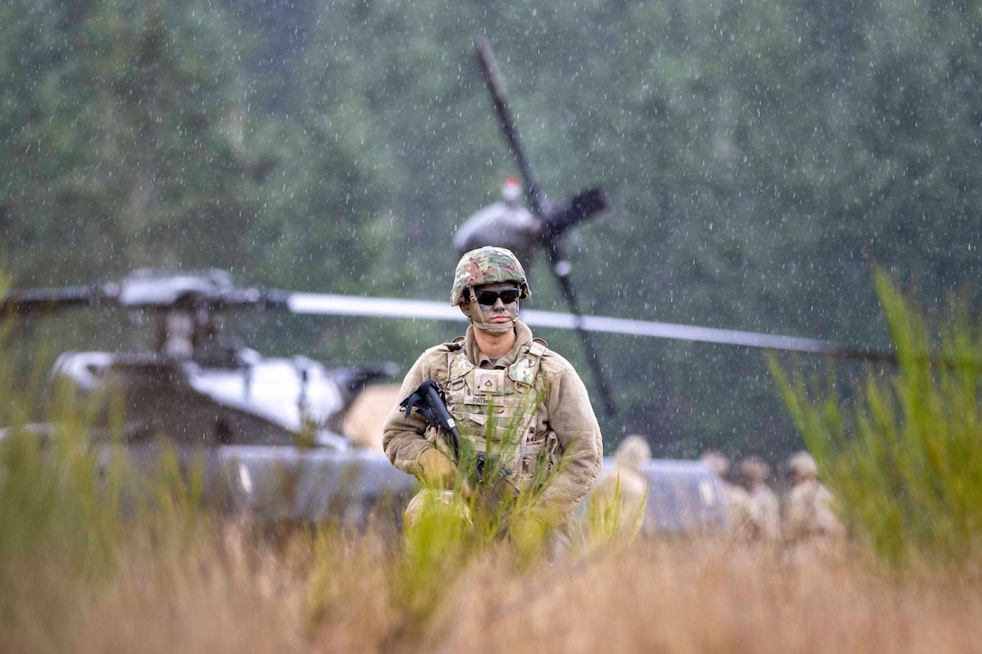 A soldier with a weapon walks through a field of tall grass with a helicopter and fellow soldiers in the background.