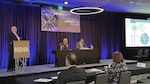 A man at a podium and two men sit at a table during a panel discussion.