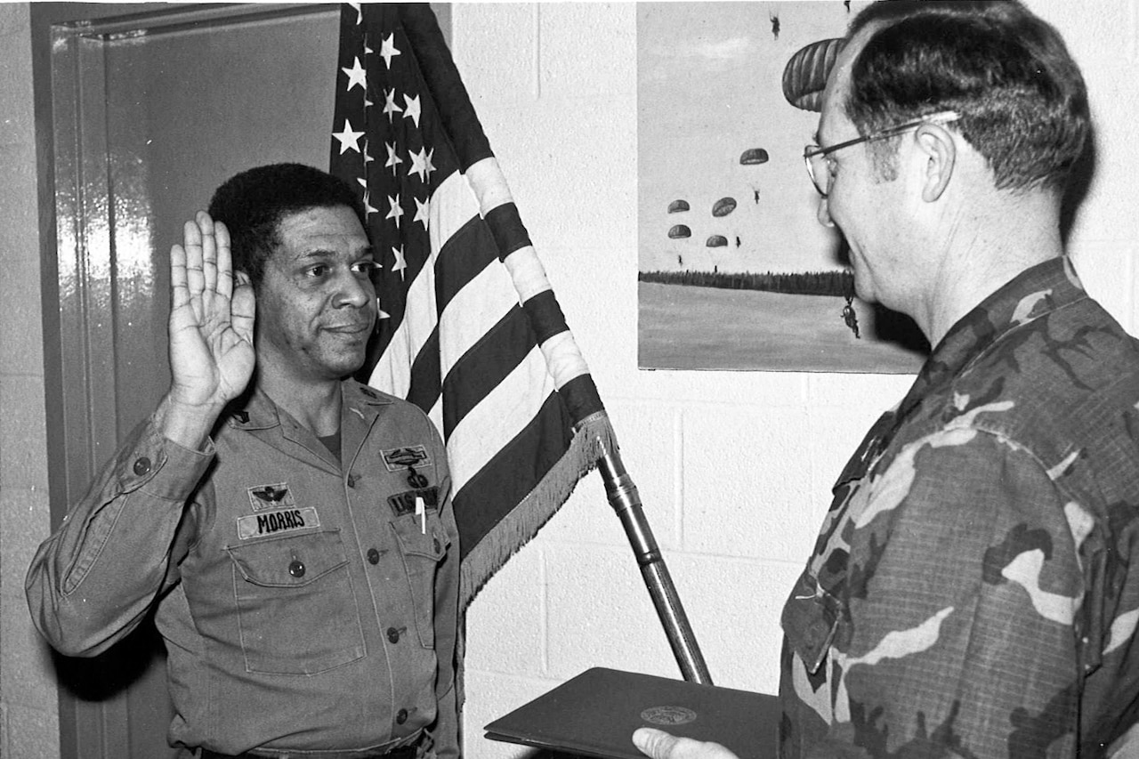 A man raises his right hand in front of another man. Both stand near an American flag.