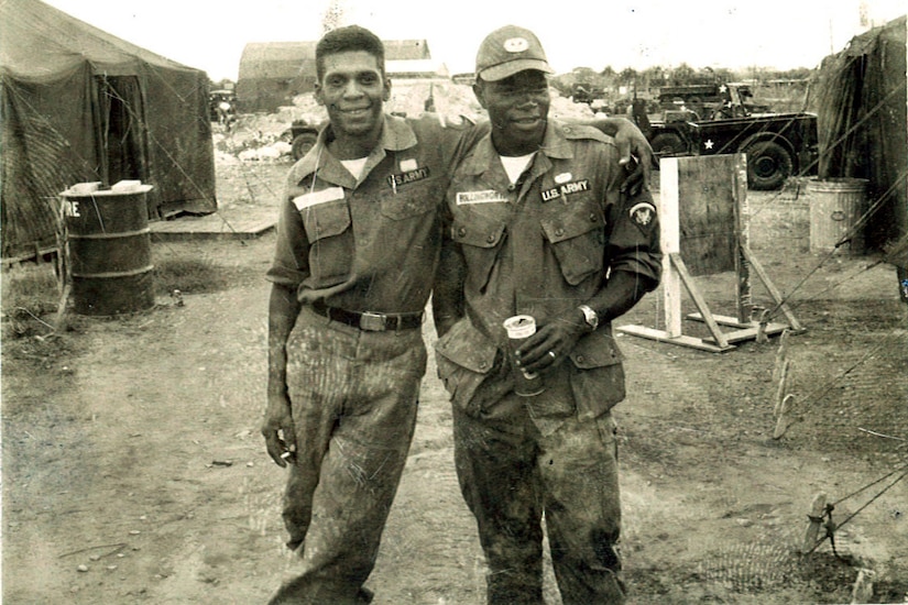 One man stands with his arm around another man’s shoulders. Tents and military vehicles are in the background.