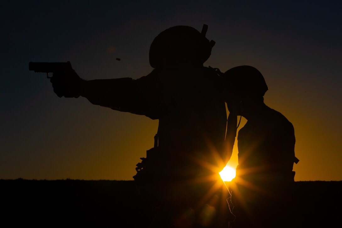 Two service members are shown in silhouette, backlit by a low sun. The soldier on the left is holding a weapon.