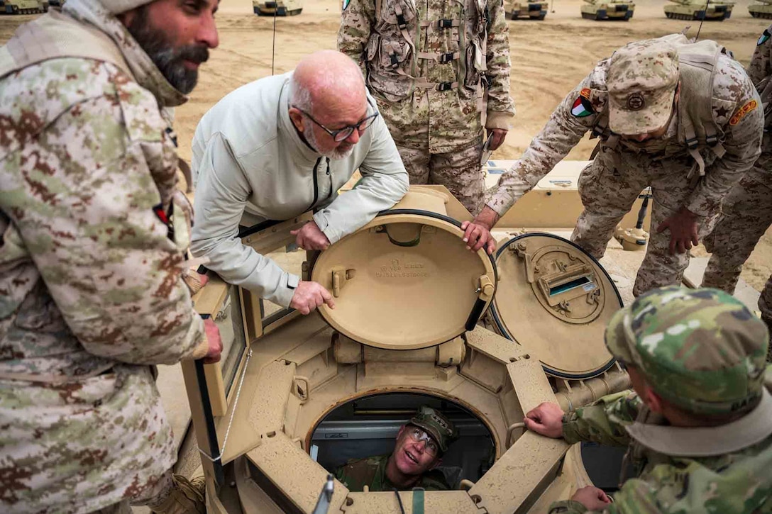 A soldier looks up from the inside of a tank as fellow service members surround the tank.