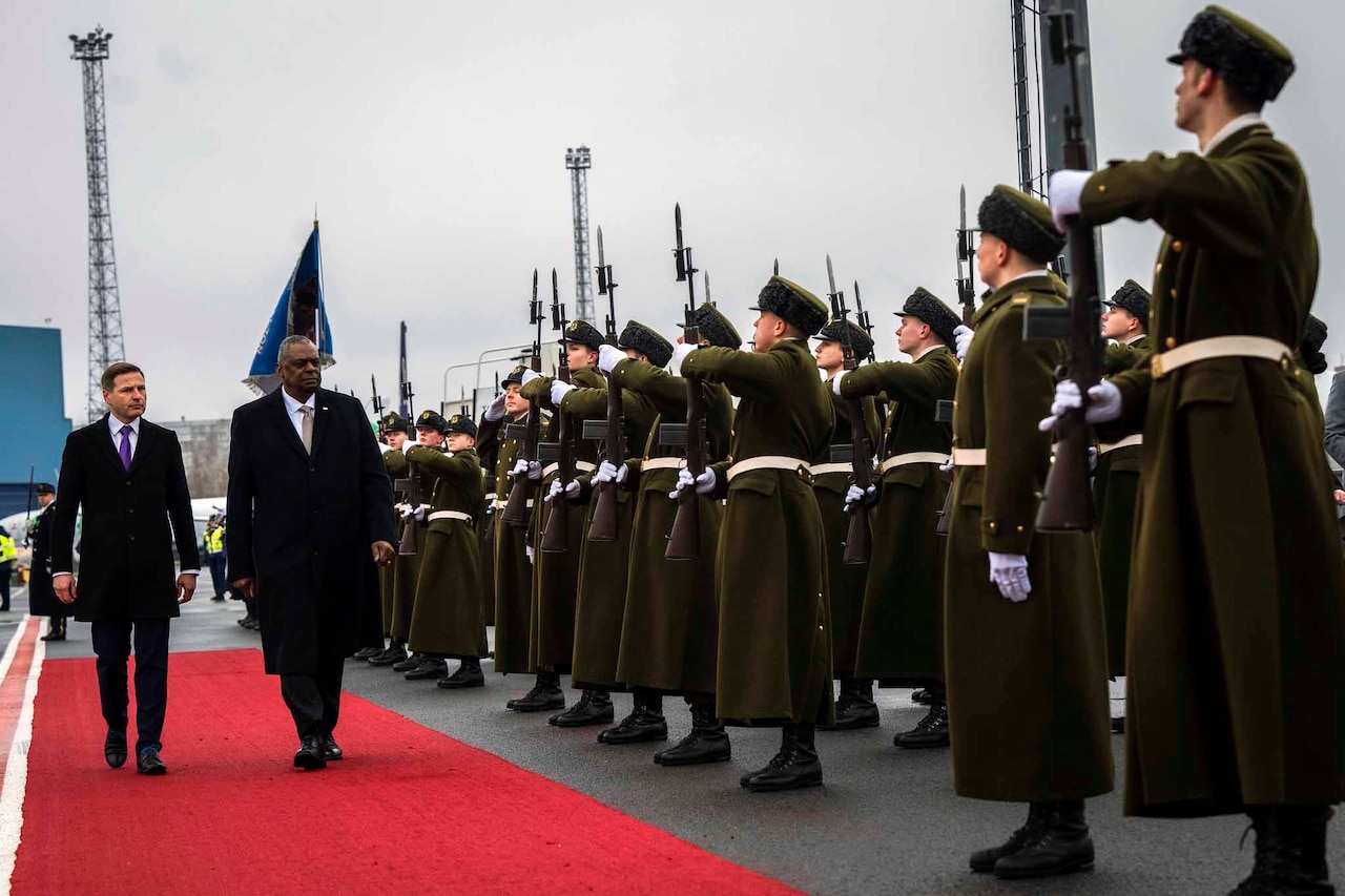 Two men walk on a red carpet in front of a group of troops.