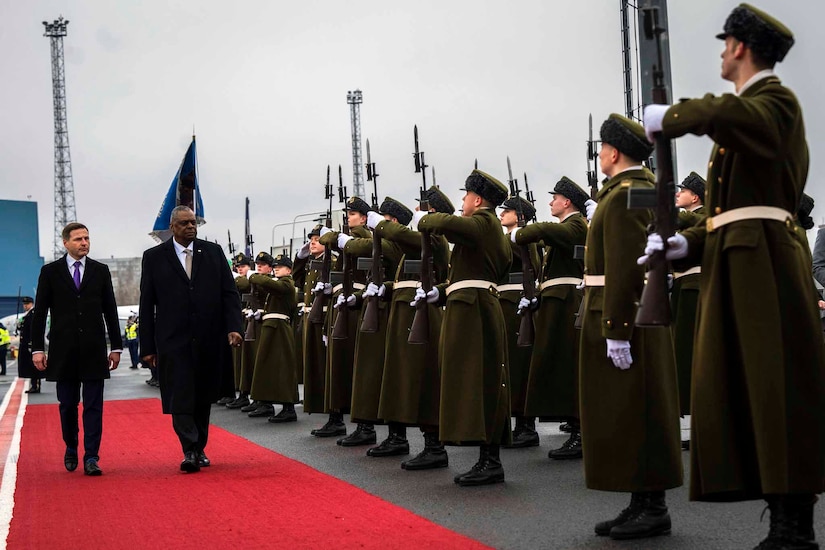 Two men walk on a red carpet in front of a group of troops.