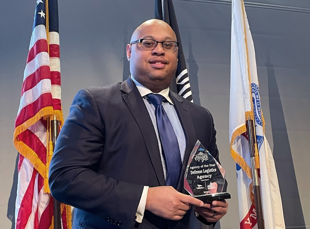 Black man in a suit stands holding a crystal plaque in front of flags