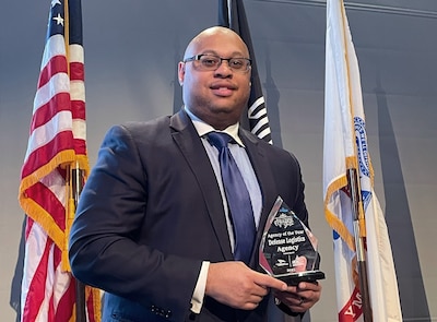 Black man in a suit stands holding a crystal plaque in front of flags