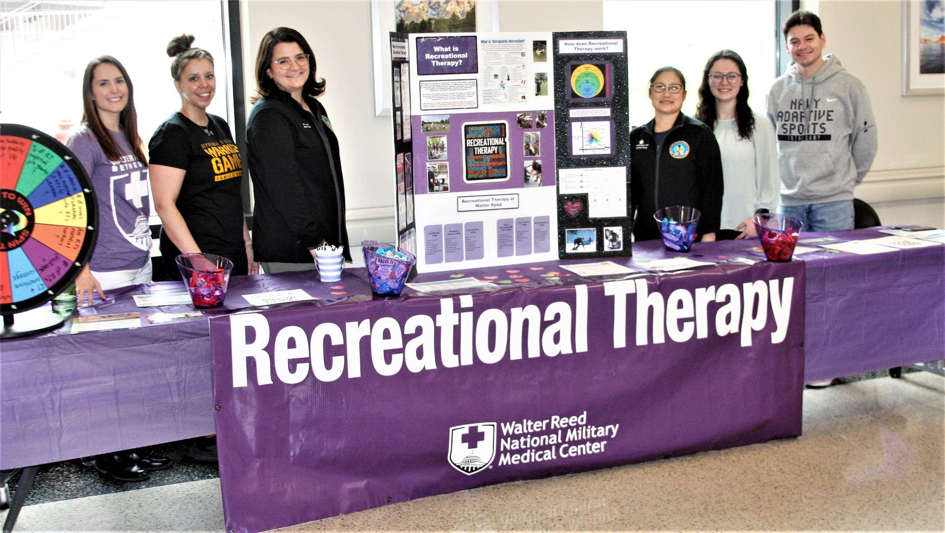 In observance of National Recreational Therapy Month celebrated during March, recreational therapists hosted an information table in the America Building lobby at Walter Reed National Military Medical Center on Feb. 16. From left, Recreational Therapists Jenny Zumwalde, Jenn Trantin, Cara Navarro, Jen Beattie, and Stephanie Seeley, along with Philip Rackham, adaptive reconditioning and sports coordinator for the Military Adaptive Sports Program (Navy), staff the table providing WRNMMC beneficiaries and staff information explaining what recreational therapy is and how it promotes the health, emotional wellbeing, skills, and abilities of people with illnesses or disabilities. Research supports the concept that people with active, satisfying lifestyles will be happier and healthier. Rec therapy is also individualized to each person by his or her past, present, and future interests, condition and lifestyle. It can used as therapy for those recovering from a stroke, rehabilitating from injury, working to improve motor skills, receiving cancer treatment, experiencing anxiety, depression or isolation, and recovering from a substance use disorder. Therapy plans use a variety of activities including arts and craft; sports and exercise; yoga and dance; music; gardening; cooking, creative writing, animal interactions and more.