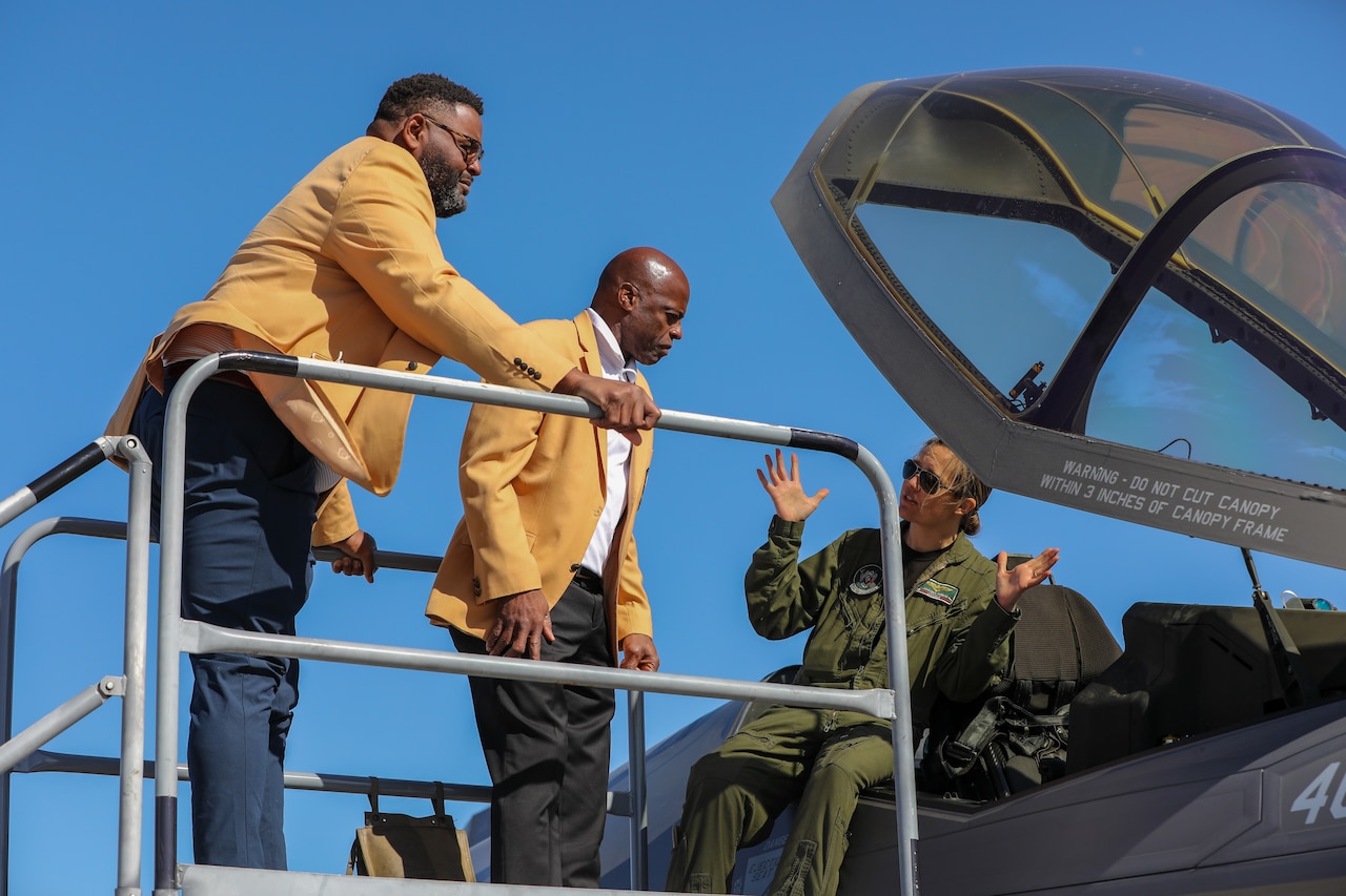 Two men wearing jackets lean over the cockpit of an aircraft as a pilot explains something to them.