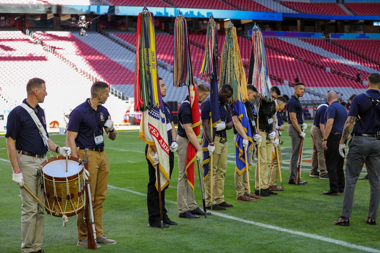 Several men holding flags, rifles and drums stand in a line as others look at their form.