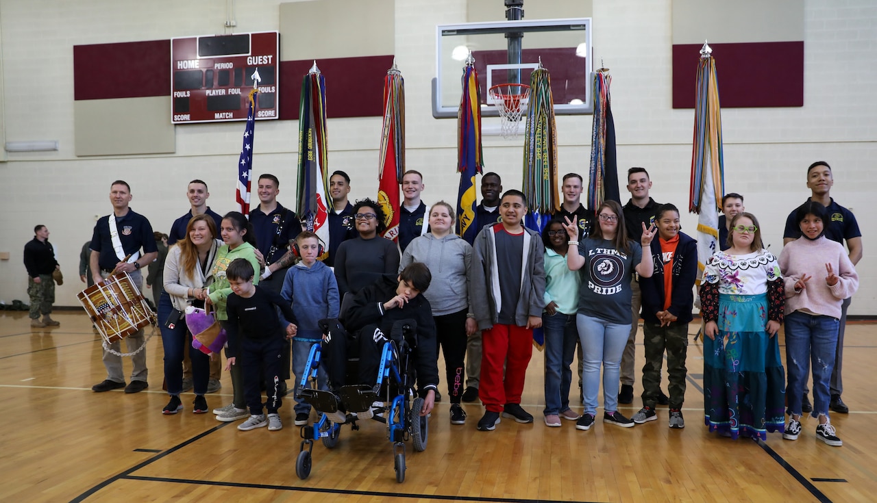 Several children stand in front of members of a color guard to pose for a photo in a gymnasium.