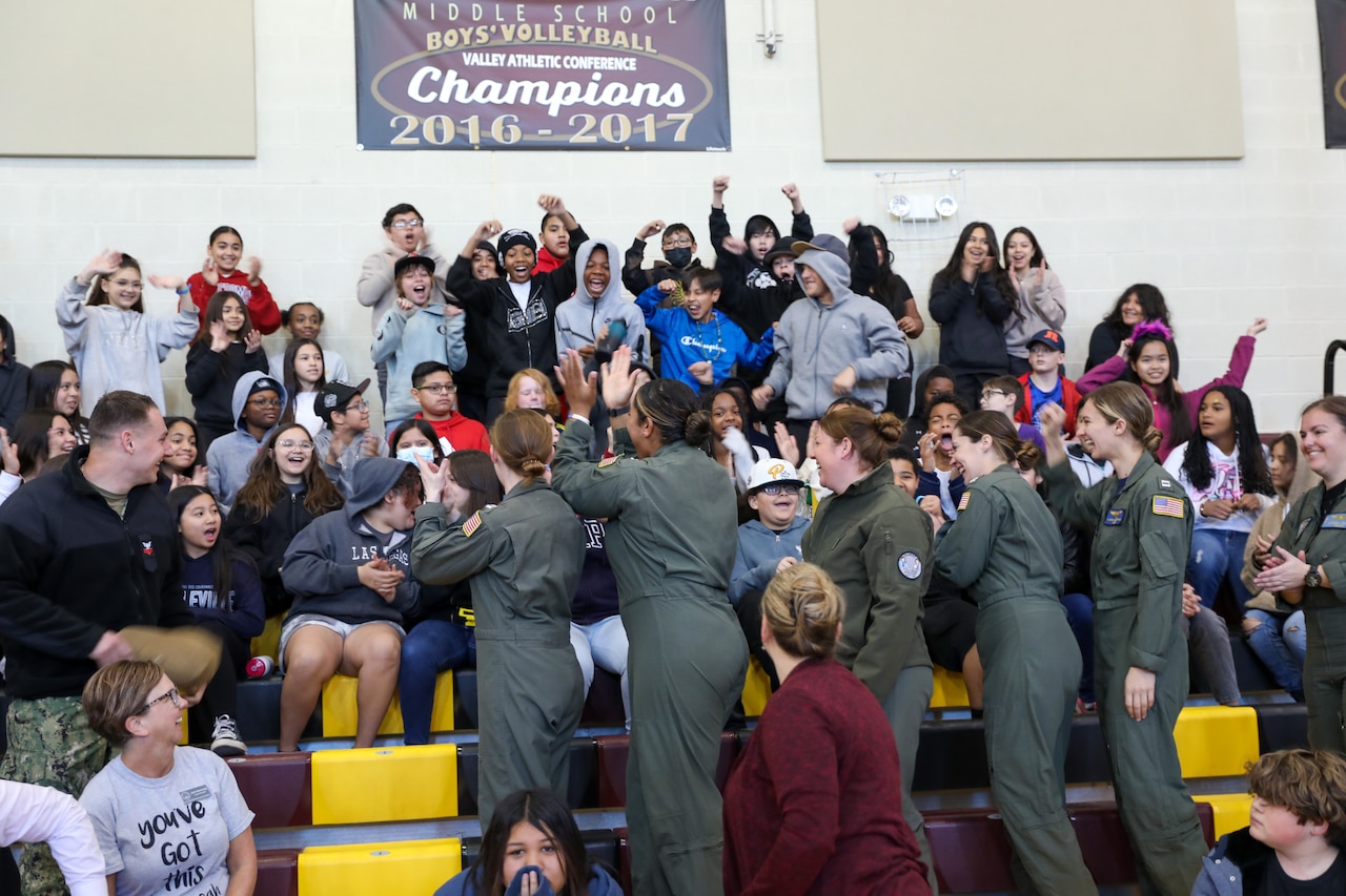 Dozens of children in bleachers cheer as five women clap in the seats in front of them.