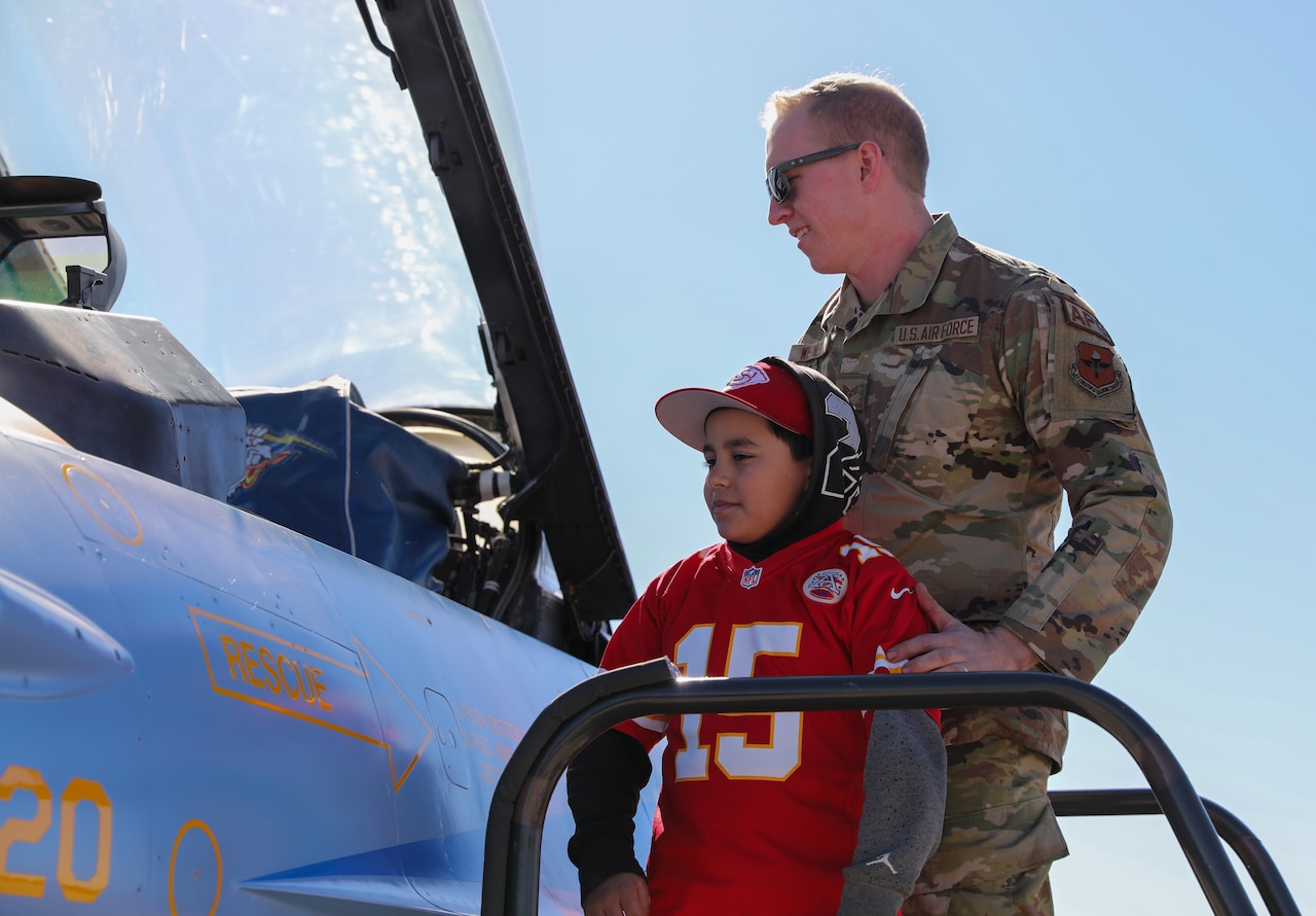 A man in camouflage uniform holds onto the arm of a young boy as they look at the cockpit of an aircraft.