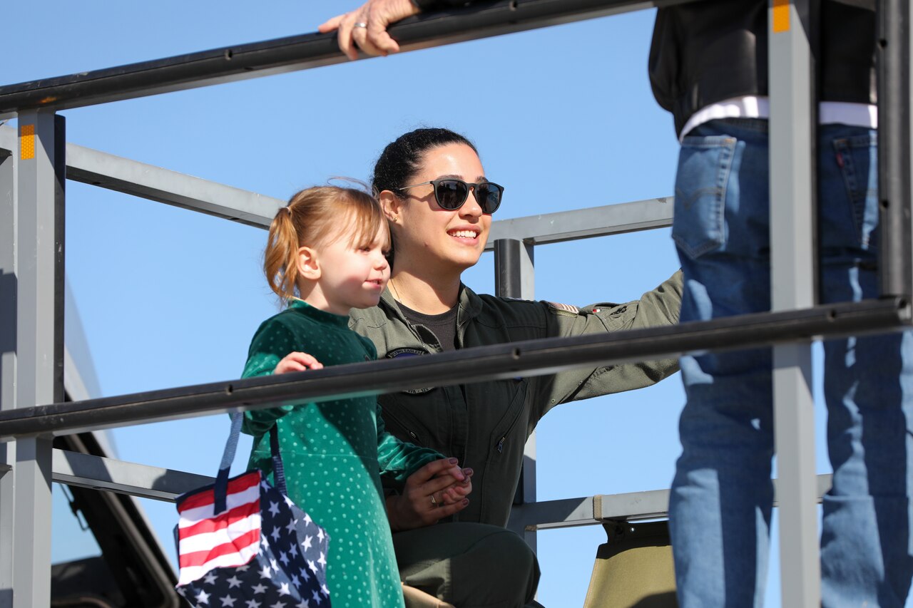 A woman in sunglasses holds hands with a young girl carrying a bag designed like a U.S. flag.