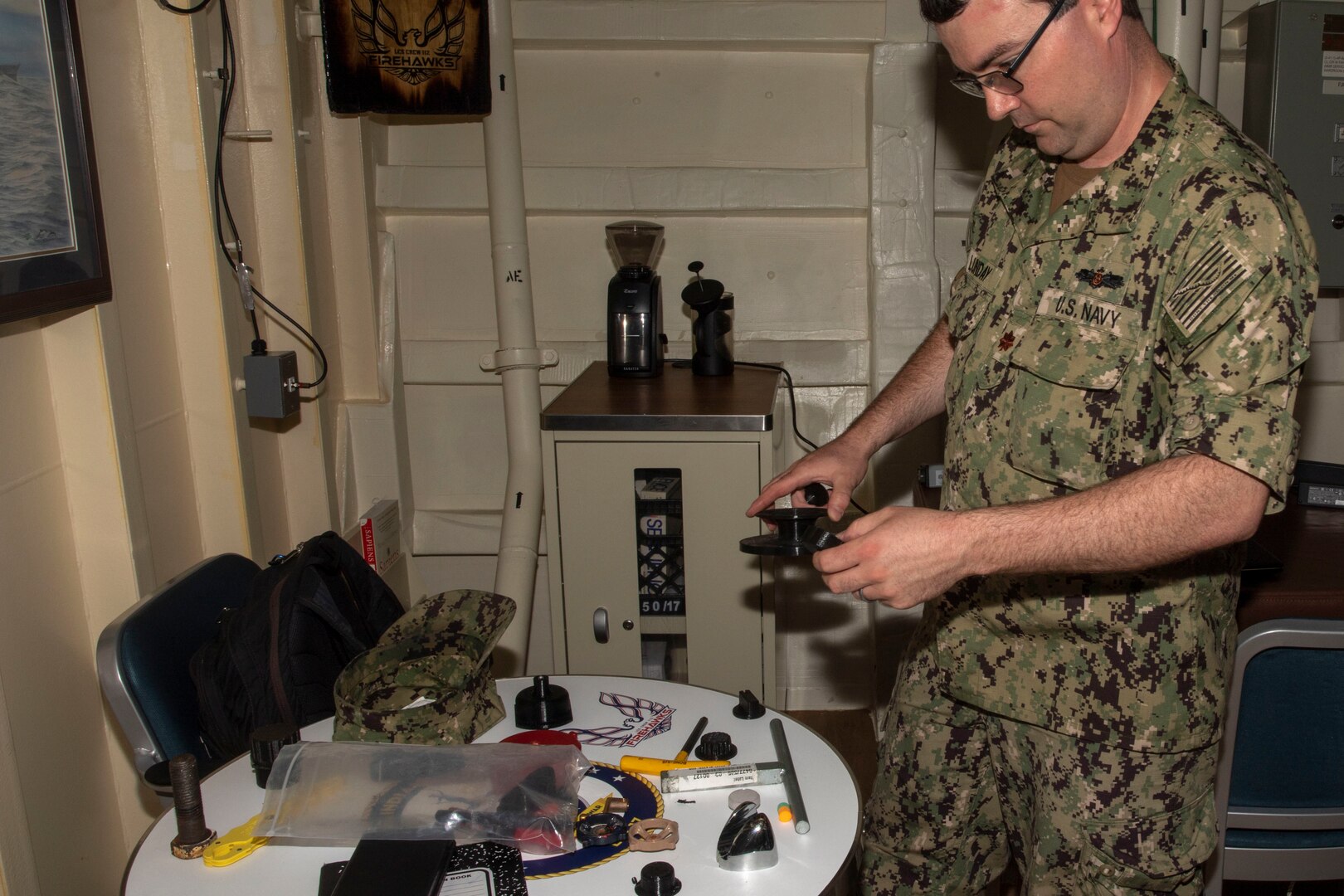 A uniformed service member holds an object while looking at a table containing various objects.