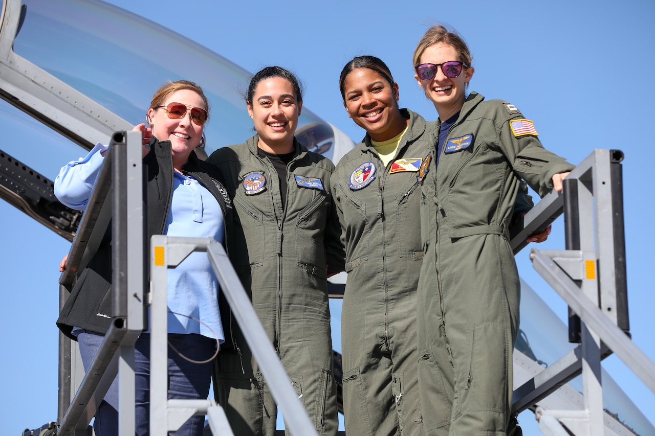 Four women, three of whom are in aviation jumpsuits, stand on a platform in front of an aircraft for a photo.