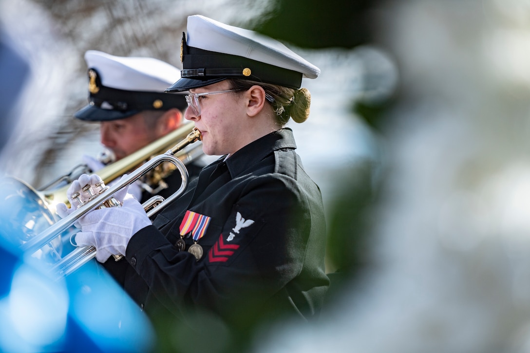 Navy musicians in formal uniforms play brass instruments at a ceremony.