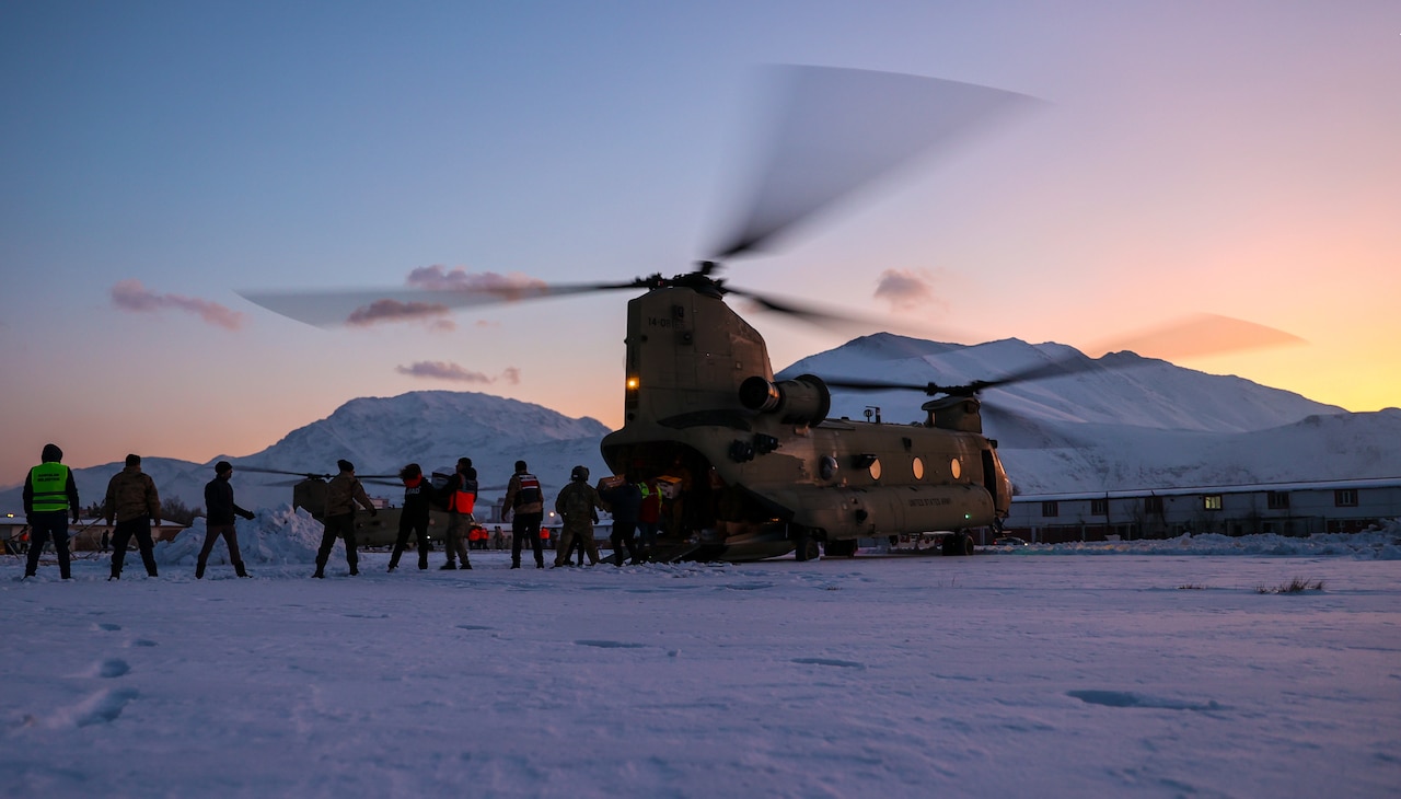 A line of soldiers and Turkish civilians offload packages from a chinook helicopter on the snow in the distance.