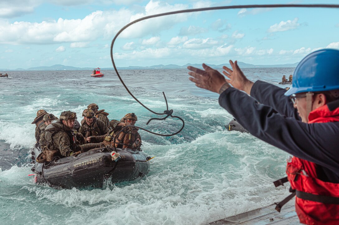 A rope is thrown between a ship and Marines in a rubber raft as the raft approaches the ship in choppy water.