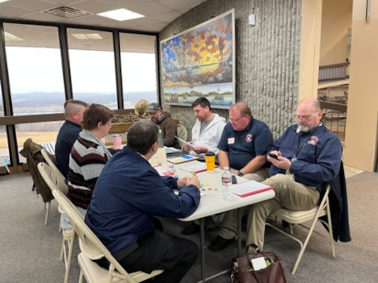 A group of USACE employees and employees from local emergency management agencies sit around a table with papers in front of them and discuss an emergency scenario.