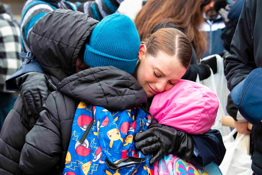 An airman hugs three children wearing black and colorful winter coats.