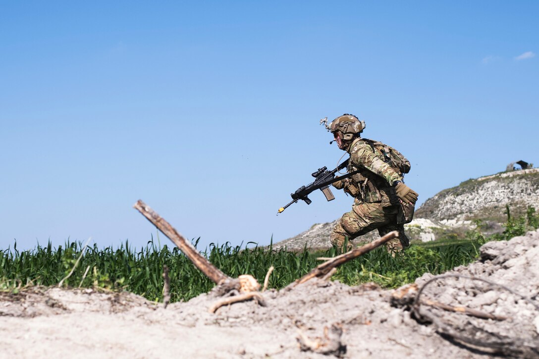 A soldier runs in a field while carrying a weapon.
