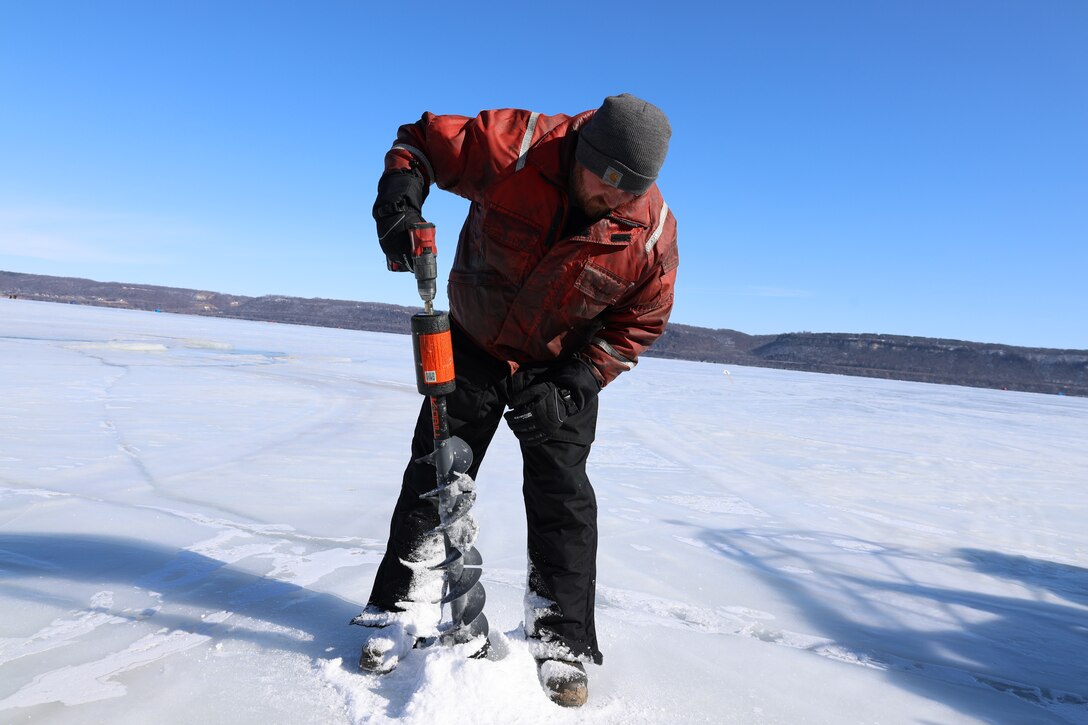 A man in winter gear holds a large drill and drills into the ice on a lake.