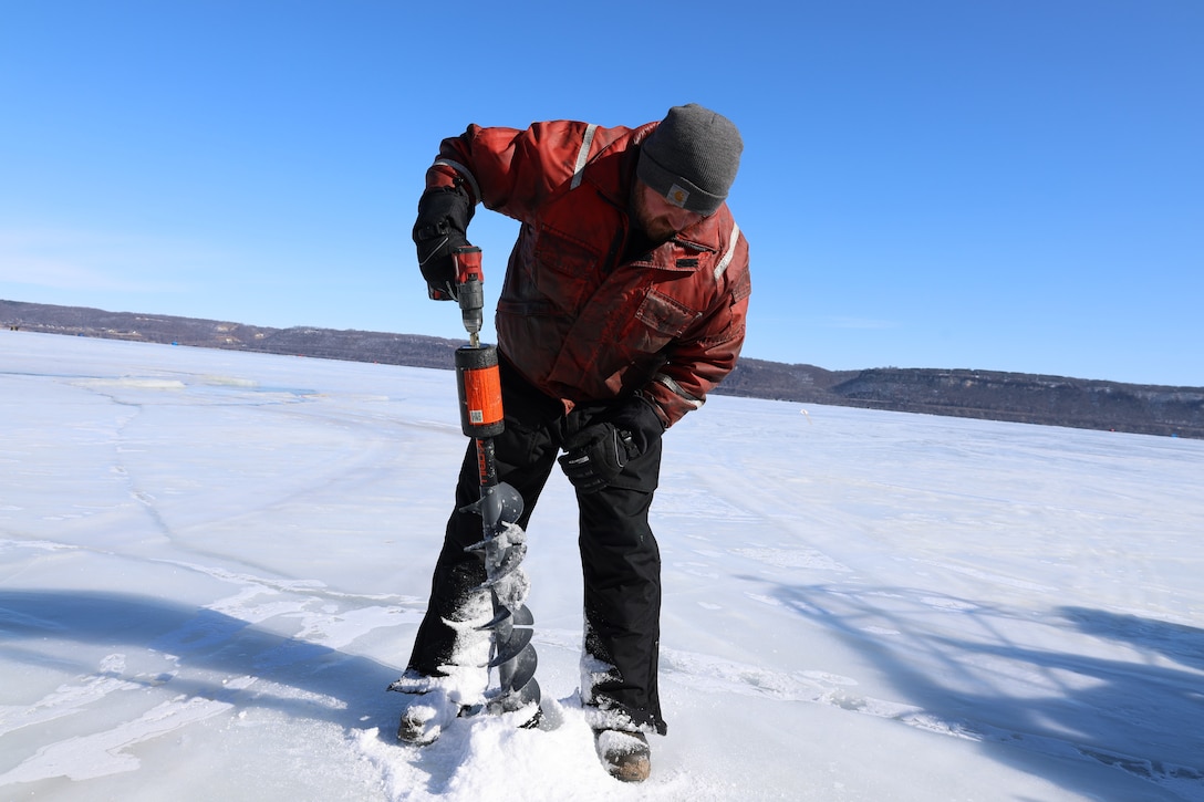 A man in winter gear holds a large drill and drills into the ice on a lake.