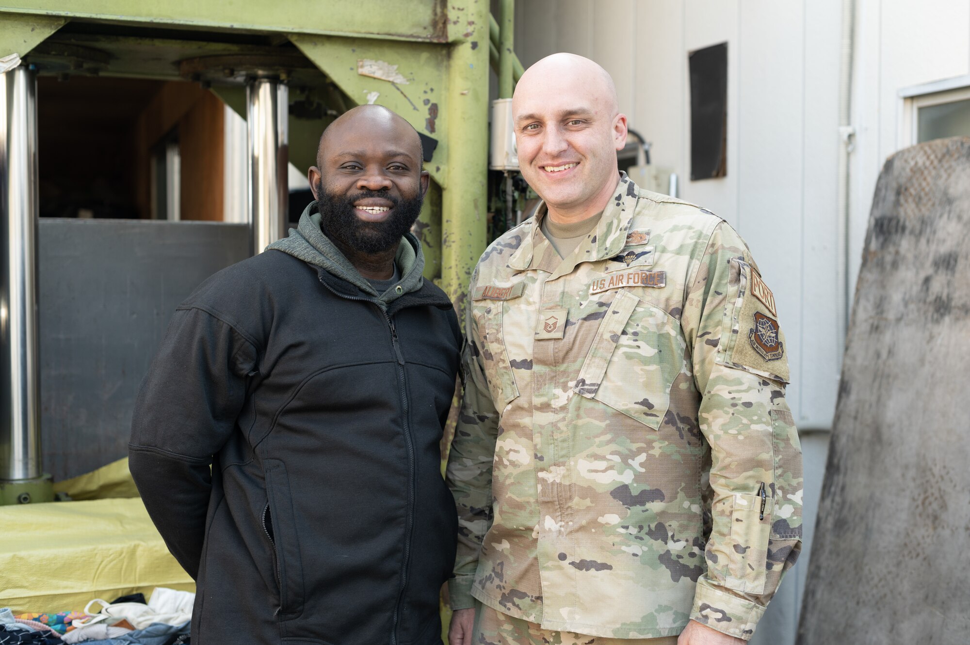 A black man and a bald white man stand next to each other while smiling for the camera.