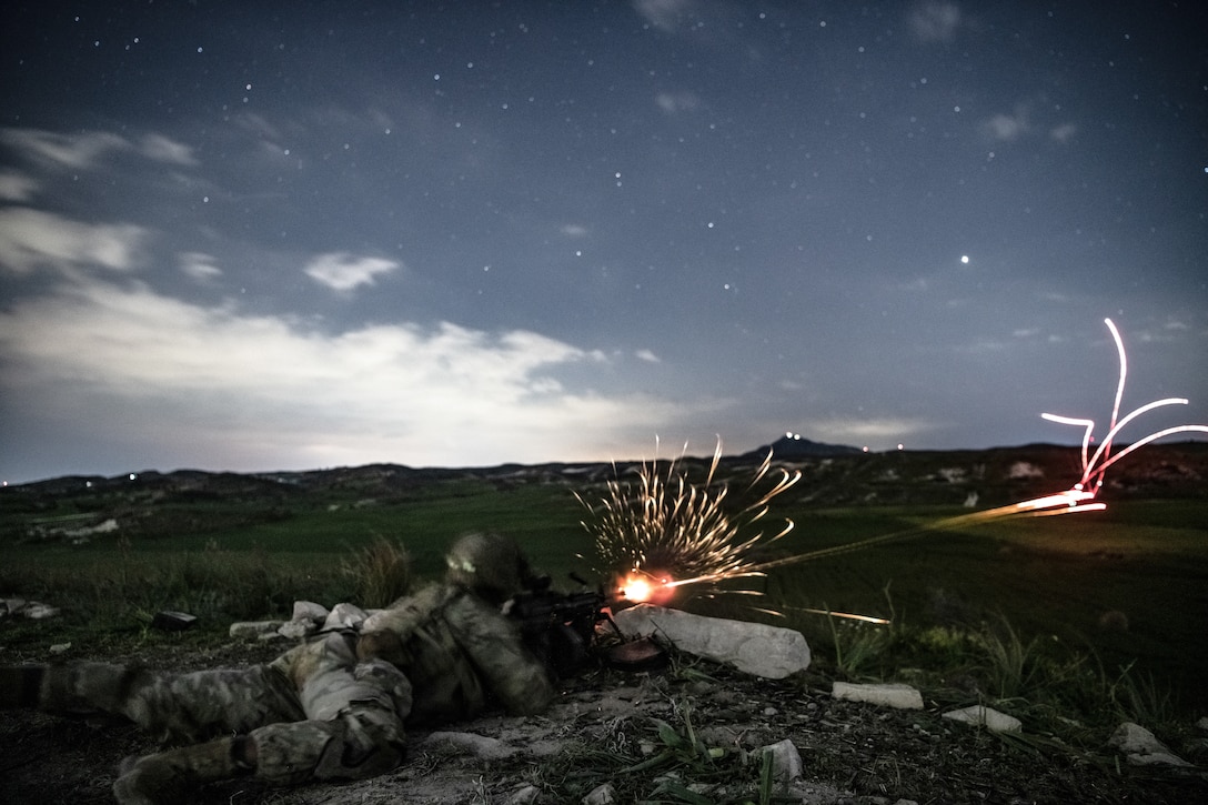 A paratrooper fires at a target while lying on the ground at dusk.