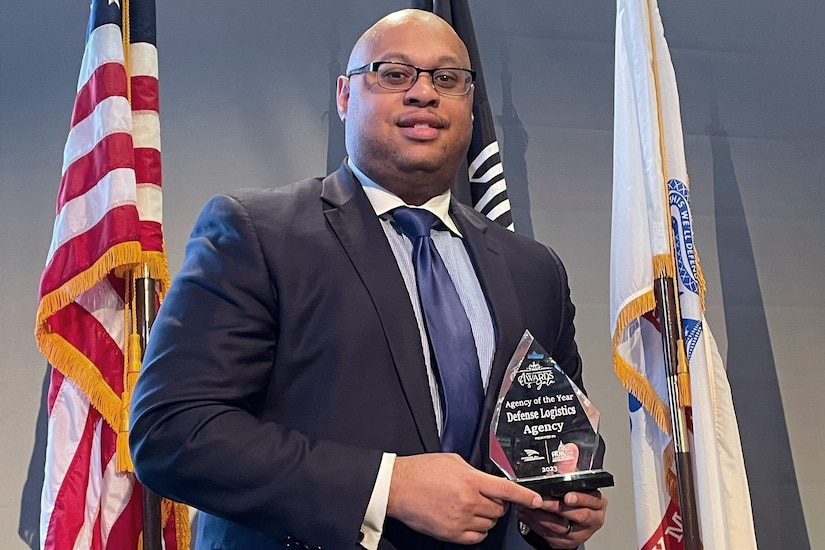 A man wearing business attire standing in front of three flags holds an award.