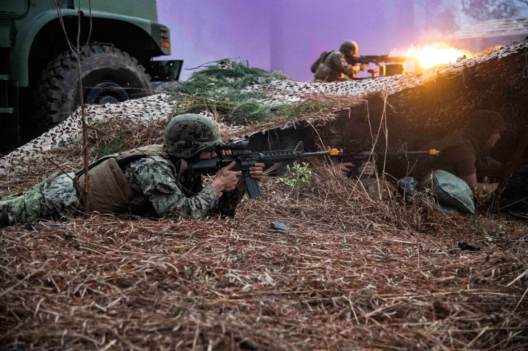 A service member fires a weapon as fellow sailors and Marines lay on the ground in camouflage aiming weapons.