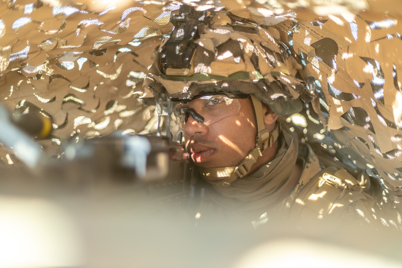 A Marine holds a weapon from underneath a tarp.